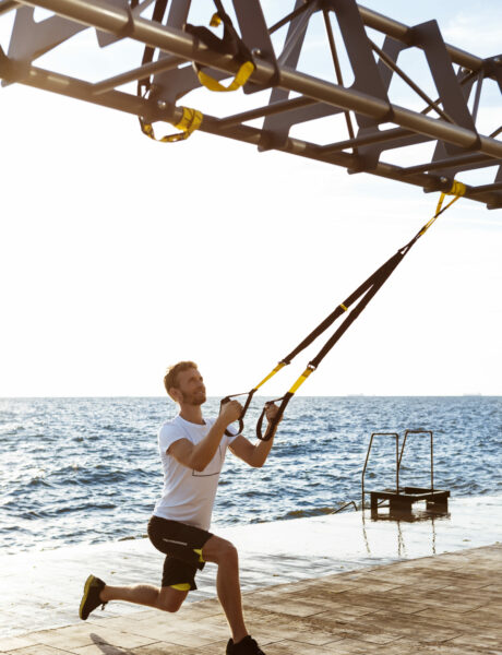 Young sportive handsome man training with trx near sea.